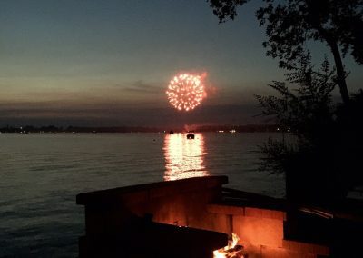Fireworks in the Sky reflecting against a lake with a fire burning in a fire pit in the foreground