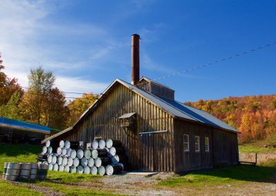 A Vermont sugar shack with a tall evaporator smokestack and stacked maple syrup barrels outside, surrounded by vibrant fall foliage on the hillside in Franklin County.