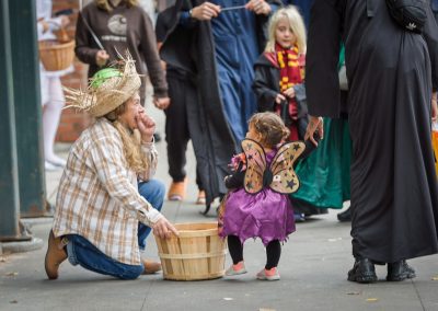 Woman dressed up as a scarecrow kneeling down offering candy to a small child in a butterfly costume